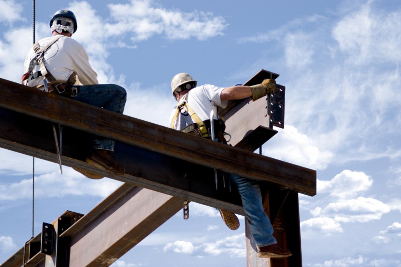 'Two ironworkers atop the skeleton of a modern building. One man is positioning a very large beam while the other watches.'