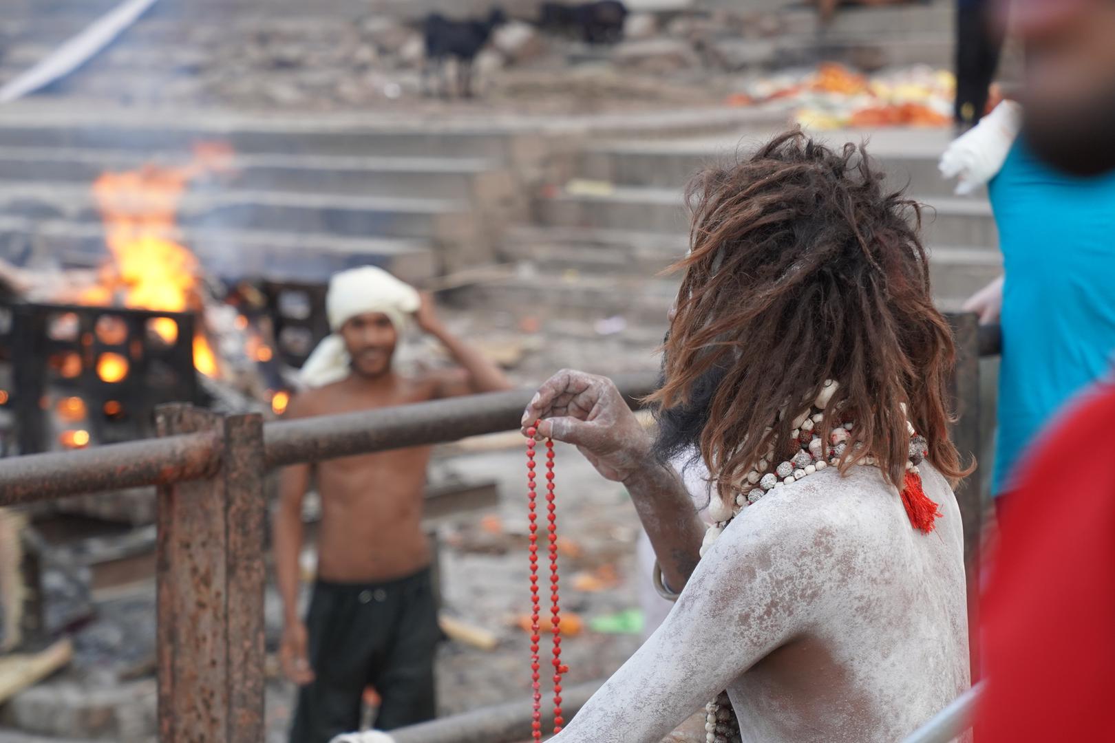 20 July 2024, India, Varanasi: A holy man prays while a corpse is burned in the background. Photo: Anne-Sophie Galli/dpa Photo: Anne-Sophie Galli/DPA