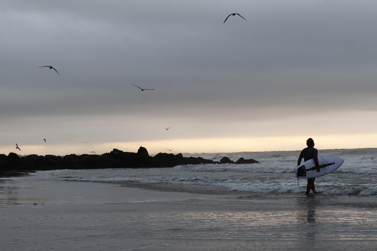 A surfer walks from the beach into the Atlantic Ocean during a cloudy sunrise in Lido Beach, New York